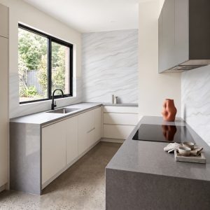 Modern kitchen with gray stone countertops, a marble backsplash, black-framed window, and minimalist cabinetry.
